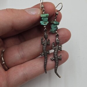 May include: Silver lizard earrings with turquoise stones. The lizards are detailed and have a textured finish. The turquoise stones are a light green color.
