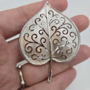 May include: A silver leaf-shaped brooch with a delicate, intricate design of swirling cutouts. The brooch is a classic and elegant piece of jewelry.
