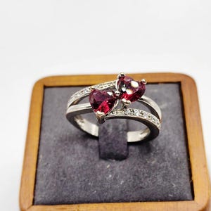 Peut inclure: Une bague argentée ornée de deux pierres précieuses rouges en forme de cœur placées côte à côte. L'anneau est divisé en deux sections, la partie supérieure étant incrustée de petites pierres claires et scintillantes sur les côtés.