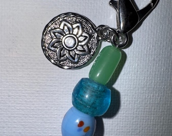 Puede incluir: Un llavero de cuentas con un mosquetón plateado y un dije decorativo. El dije es un colgante circular plateado con un diseño floral. Las cuentas son de varios colores, incluyendo azul, verde y transparente.