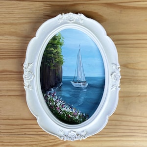 Puede incluir: Pintura ovalada en un marco blanco ornamentado. La obra representa un velero sobre agua azul, con un acantilado y árboles a la izquierda. Flores bordean la orilla del agua. El marco tiene detalles florales decorativos.