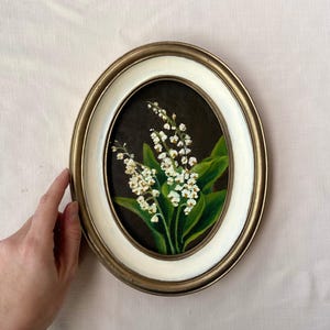 May include: Oval framed painting of lily of the valley flowers. The artwork features white flowers with green leaves against a dark background. The frame is gold and white, with a hand holding the frame.