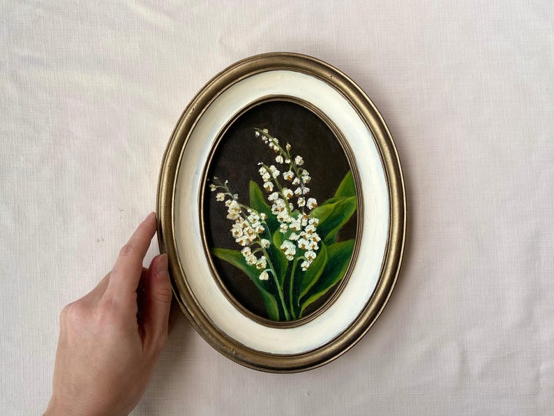 May include: Oval framed painting of lily of the valley flowers. The artwork features white flowers with green leaves against a dark background. The frame is gold and white, with a hand holding the frame.