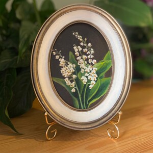 May include: Oval framed painting of lily of the valley flowers. The artwork features white flowers with green leaves against a dark background. The frame is gold and white, and the painting is displayed on a gold easel.