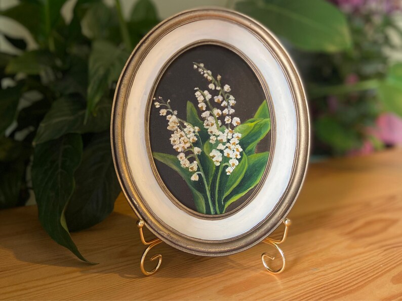 May include: Oval framed painting of lily of the valley flowers. The artwork features white flowers with green leaves against a dark background. The frame is gold and white, and the painting is displayed on a gold easel.
