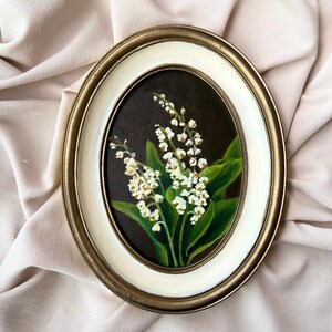 May include: Oval framed painting of lily of the valley flowers. The artwork features white blossoms and green leaves against a dark background. The frame has a gold and white border, adding a classic touch to the floral art.