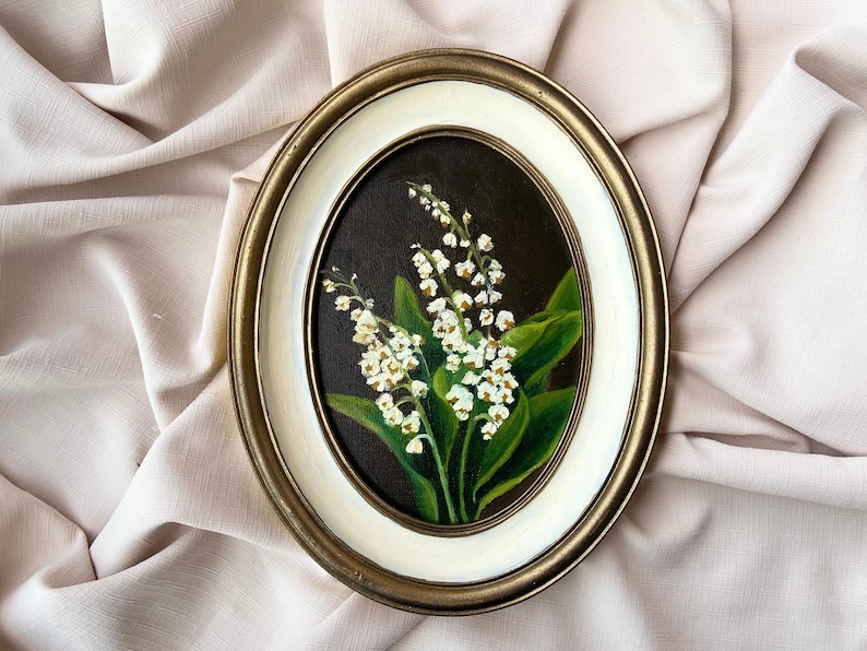 May include: Oval framed painting of lily of the valley flowers. The artwork features white blossoms and green leaves against a dark background. The frame has a gold and white border, adding a classic touch to the floral art.