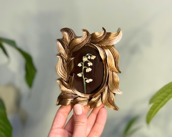 May include: An ornate gold-coloured frame with a dark brown background and a single sprig of white lily of the valley flowers.