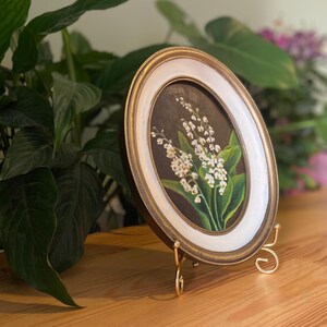 May include: Oval framed painting of lily of the valley flowers. The artwork features white flowers with green leaves against a dark background. The frame is gold with a white inner border. The painting is displayed on a gold easel.