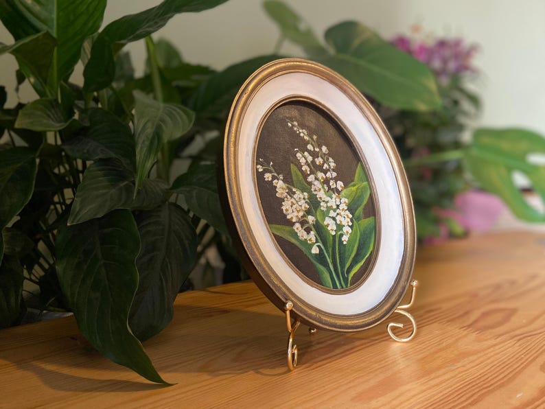 May include: Oval framed painting of lily of the valley flowers. The artwork features white flowers with green leaves against a dark background. The frame is gold with a white inner border. The painting is displayed on a gold easel.