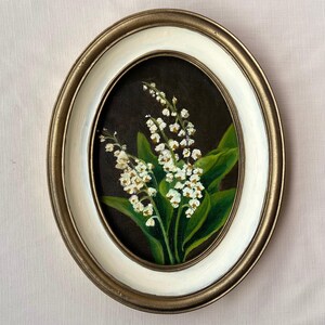 May include: Oval framed painting of lily of the valley flowers. The artwork features white flowers with green leaves against a dark background. The frame is gold with a white inner border, creating a classic aesthetic.