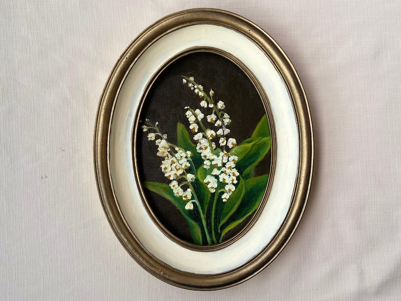 May include: Oval framed painting of lily of the valley flowers. The artwork features white flowers with green leaves against a dark background. The frame is gold with a white inner border, creating a classic aesthetic.