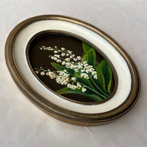 May include: An oval framed painting depicting lily of the valley. The artwork shows white flowers and green leaves set against a dark backdrop. The frame has a gold-tone outer edge and a white inner border.