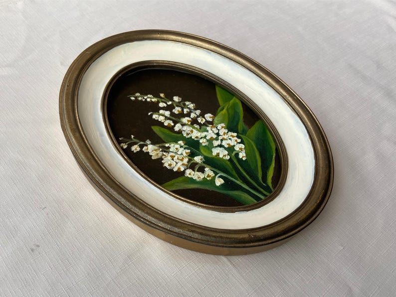 May include: An oval framed painting depicting lily of the valley. The artwork shows white flowers and green leaves set against a dark backdrop. The frame has a gold-tone outer edge and a white inner border.