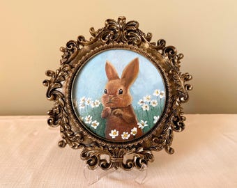 May include: An oval painting of a brown bunny in a field of white flowers, set in an ornate, gold-colored frame. The artwork is displayed on a white surface, creating a charming, vintage aesthetic. The frame is approximately 8 inches wide.