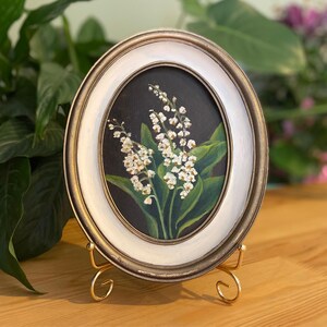 May include: Oval framed painting of lily of the valley flowers. The artwork features white flowers with green leaves against a dark background. The frame is white with a gold trim and is displayed on a gold stand.