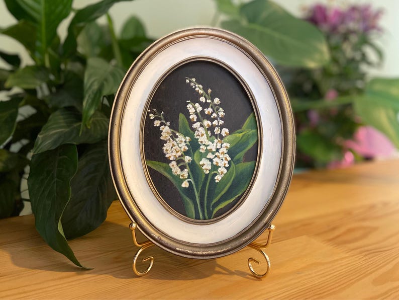May include: Oval framed painting of lily of the valley flowers. The artwork features white flowers with green leaves against a dark background. The frame is white with a gold trim and is displayed on a gold stand.