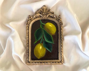 May include: A framed painting of two yellow lemons on a branch with green leaves. The artwork is set against a dark brown background and is enclosed in an ornate, gold-coloured frame. The painting is displayed on a white, textured fabric.
