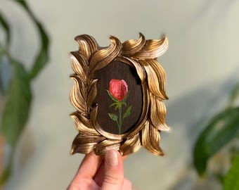 May include: A single red rose painted on canvas is framed in a gold ornate frame. The frame is made of a series of overlapping leaves.