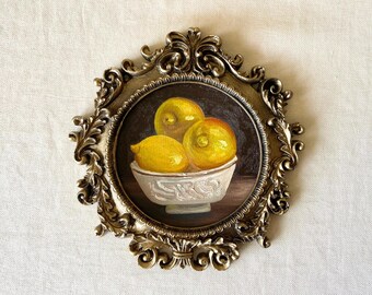 May include: A small, framed oil painting depicting three lemons in a white bowl. The painting is set against a dark background and framed in an ornate, gold-coloured oval frame. The lemons are yellow and the bowl is white.