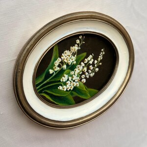 May include: Oval framed painting of white lily of the valley flowers with green leaves on a dark backdrop. The frame has a gold-tone outer edge and a white inner edge.