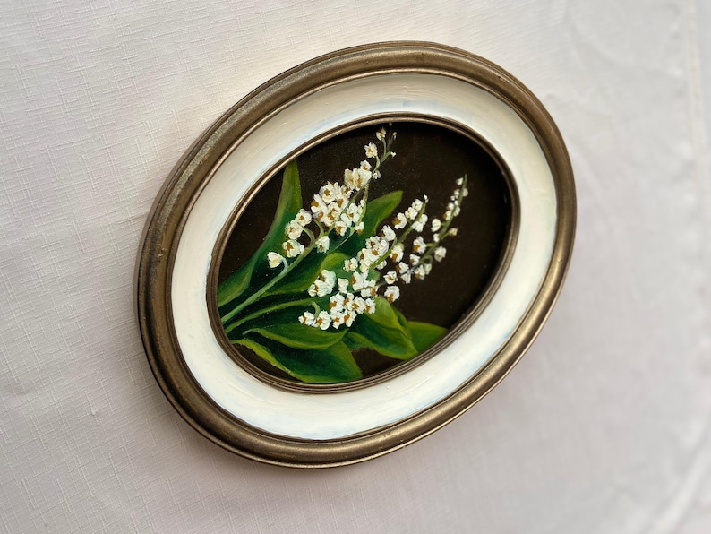May include: Oval framed painting of white lily of the valley flowers with green leaves on a dark backdrop. The frame has a gold-tone outer edge and a white inner edge.