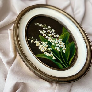 May include: An oval framed painting of lily of the valley. The artwork depicts white flowers and green leaves on a dark background. The frame has a gold-tone outer edge and a white inner border.