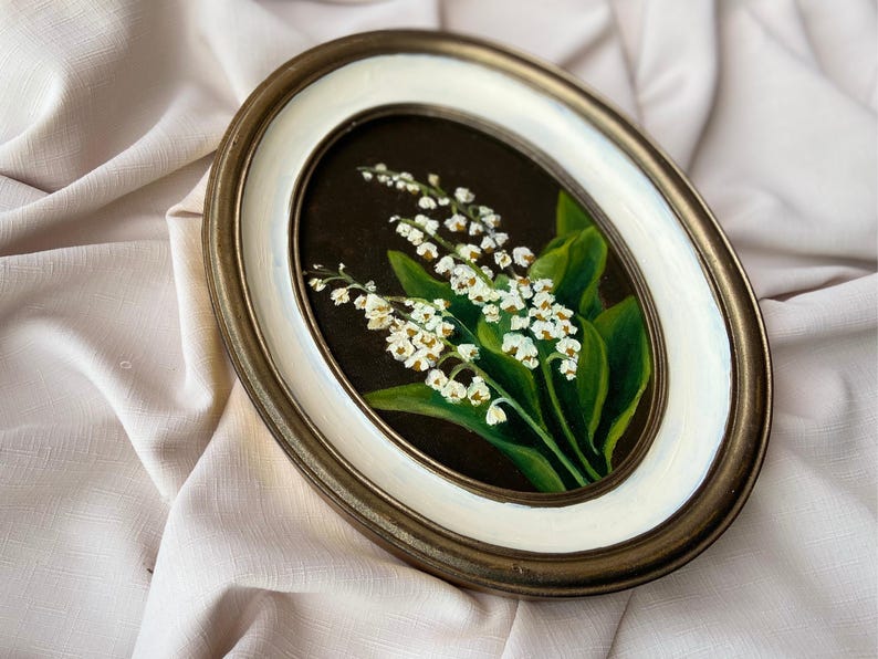 May include: An oval framed painting of lily of the valley. The artwork depicts white flowers and green leaves on a dark background. The frame has a gold-tone outer edge and a white inner border.