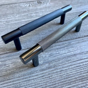 May include: Two black metal cabinet pulls with a textured finish. The pulls have a T-shaped design.