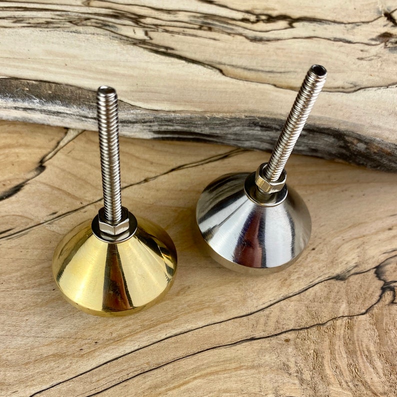 May include: Two metal furniture feet, one gold and one silver, with a threaded screw for attaching to furniture. The feet are on a wooden surface.
