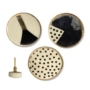 May include: A set of three ceramic cabinet knobs with gold edging. The knobs display black and white geometric patterns, including a pie chart and polka dots. One knob is shown with its brass fitting.