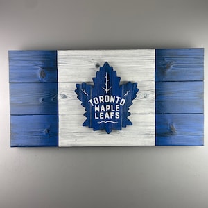 May include: A blue and white wooden sign with the Toronto Maple Leafs logo. The logo is a blue maple leaf with the words "Toronto Maple Leafs" written in white inside the leaf.