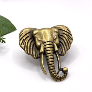 May include: A gold-toned elephant head brooch with detailed features. The elephant's trunk is curled up and the ears are large and fan-shaped.