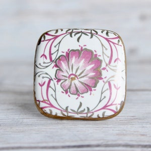 May include: Square ceramic cabinet knob with a white background and a pink floral design. The knob has a gold trim.
