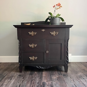 May include: A dark brown wooden cabinet featuring three horizontal drawers with ornate gold-toned metal pulls, one side door with a small round knob, and carved leaf-like detailing on the front vertical edges, topped with a potted orchid.