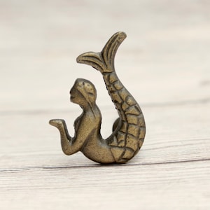 May include: A gold-toned mermaid figurine with a detailed tail and a sitting pose. The mermaid is holding her hand up to her face.