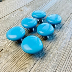 May include: Six light blue ceramic cabinet knobs with silver metal bases. The knobs are arranged in a semi-circle on a light wood surface.