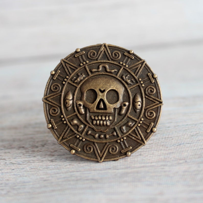 Skull Drawer Knob in Antique Brass. The knob is round and made of metal. It looks like a pirate doubloon.