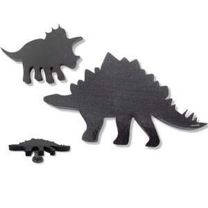 Peut inclure: Trois silhouettes de dinosaures en bois noir. Le plus grand dinosaure est un stégosaure, le deuxième plus grand est un tricératops et le plus petit est un stégosaure plus petit.