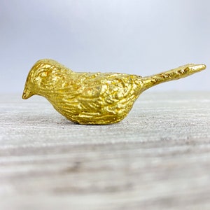May include: A small, gold-coloured bird figurine with a textured surface. The bird is perched on a light wood surface.