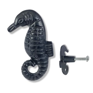 May include: Black metal seahorse door handle with a decorative design. The handle is attached to a metal plate with a screw.