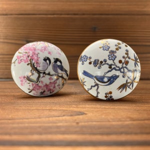 May include: Two round ceramic cabinet knobs with bird and floral designs. One knob features pink cherry blossoms and two birds. The other knob has a blue bird and blue floral branches with gold accents. Both knobs have gold rims.