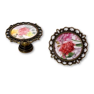May include: Two antique bronze cabinet knobs with floral designs. One knob is mounted on a short pedestal. The floral designs feature pink, red, and yellow flowers with green leaves on a white background. The knobs have a decorative, openwork border.