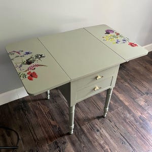 May include: A sage green drop-leaf table featuring two drawers with gold-toned bird-shaped pulls. The tabletop surfaces on the left and right sides are decorated with colourful floral and butterfly decals, while the legs are turned and tapered.