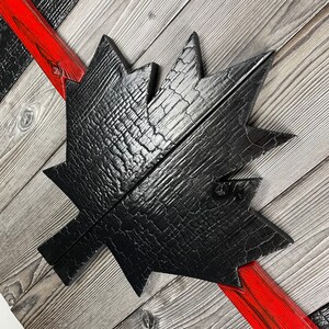 May include: A black wooden maple leaf with a distressed, charred finish. The leaf is mounted on a wooden background with red painted stripes.