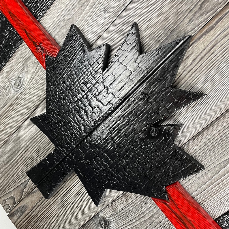 May include: A black wooden maple leaf with a distressed, charred finish. The leaf is mounted on a wooden background with red painted stripes.