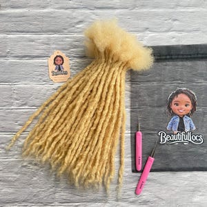 May include: Blonde dreadlocks arranged in a bundle, with two pink crochet hooks and a tag featuring a cartoon illustration. The tag and sticker both display the text "Beautifullocs". The dreadlocks are a light, golden color.