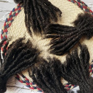 May include: Several bundles of dark brown dreadlocks are arranged on a round, woven trivet. The trivet has a cream-colored center and a red, white, and blue braided border. The dreadlocks are tightly coiled and appear to be made of natural hair.