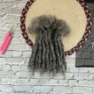 May include: A bundle of gray dreadlocks rests on a beige, round placemat with a red, white, and blue braided border. Two pink-handled crochet hooks are visible on the left side of the image. The dreadlocks appear to be made of a textured material.