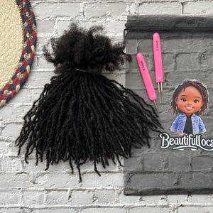 May include: A bundle of black dreadlocks with a textured appearance. Two pink crochet hooks are next to the dreadlocks. A sticker with the text "Beautifullocs" and a cartoon doll is also in the image.